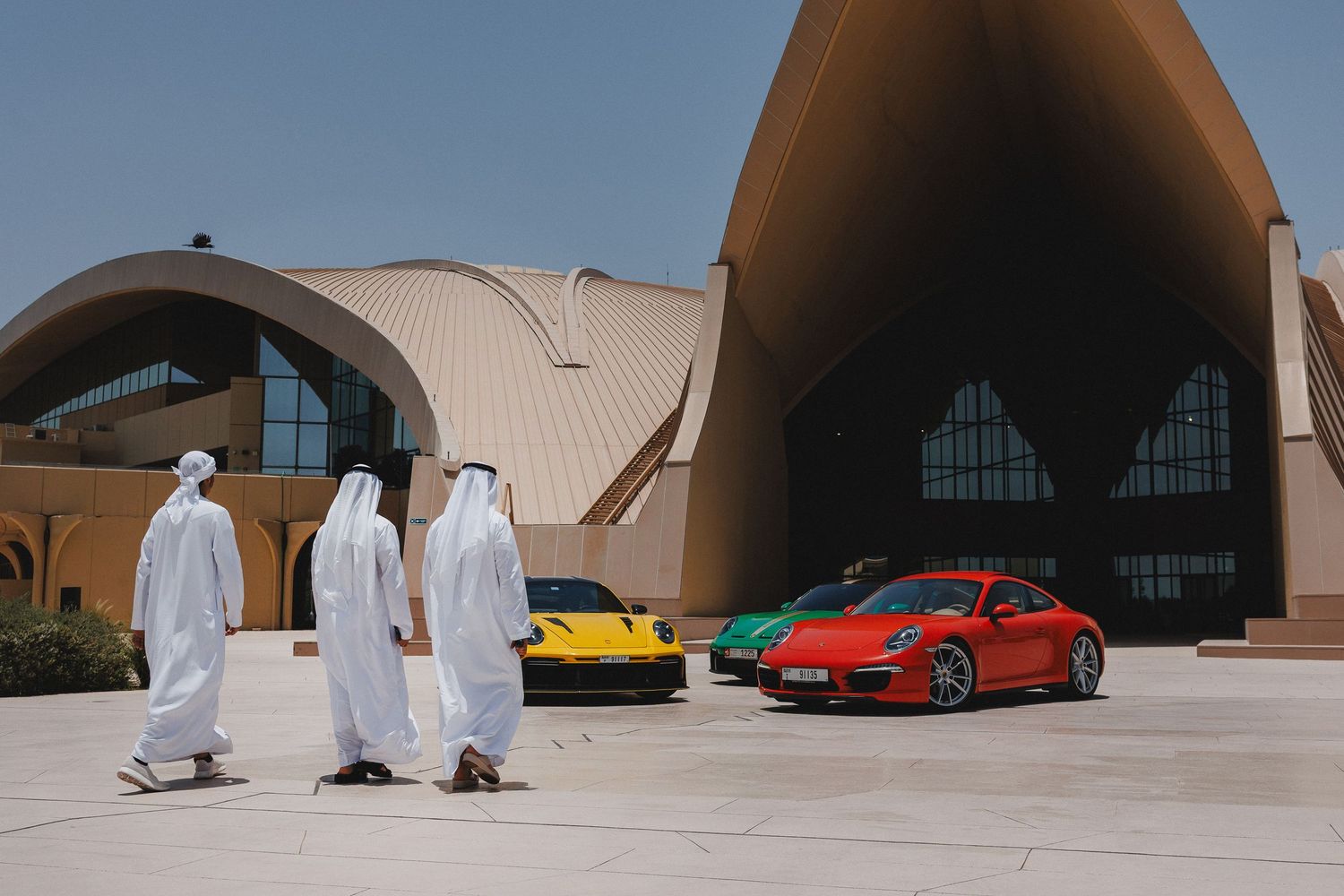 Retro cars displayed in front of curved modernist architecture in desert setting.