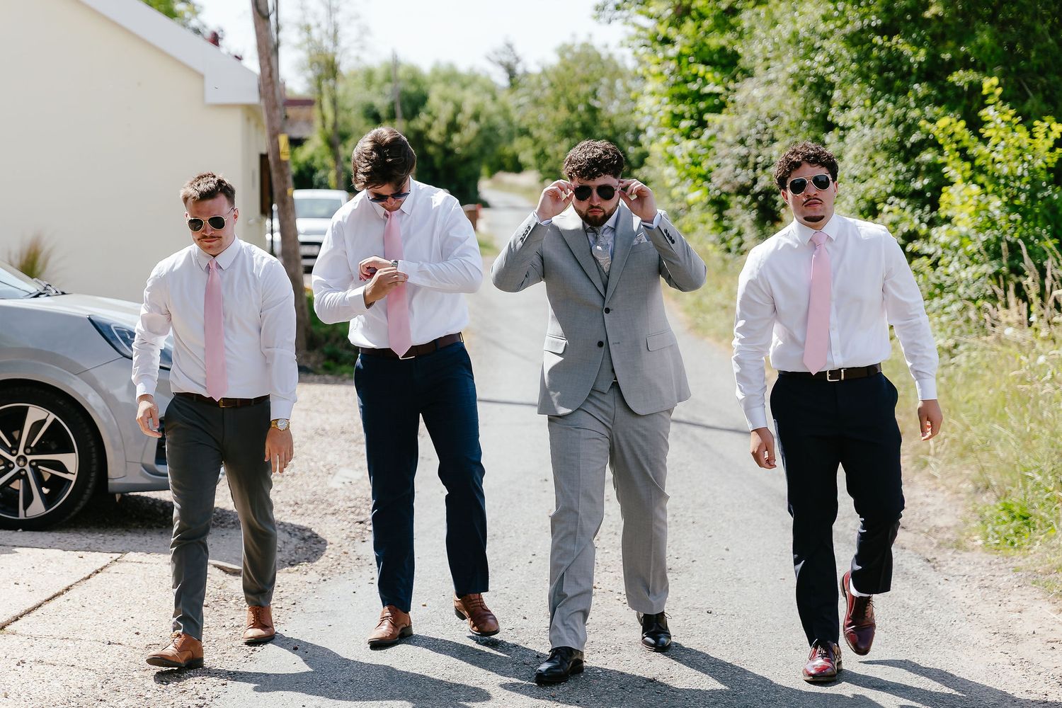 Four groomsmen in pink ties and white shirts walk together outdoors on a sunny wedding day.