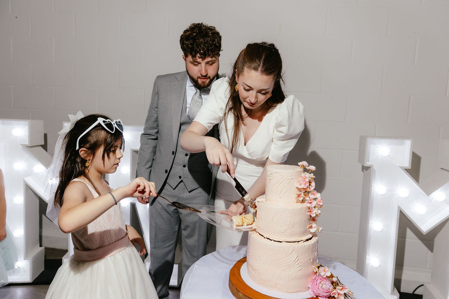 Bride and groom cutting a pink floral wedding cake with a young flower girl in a white dress beside them.