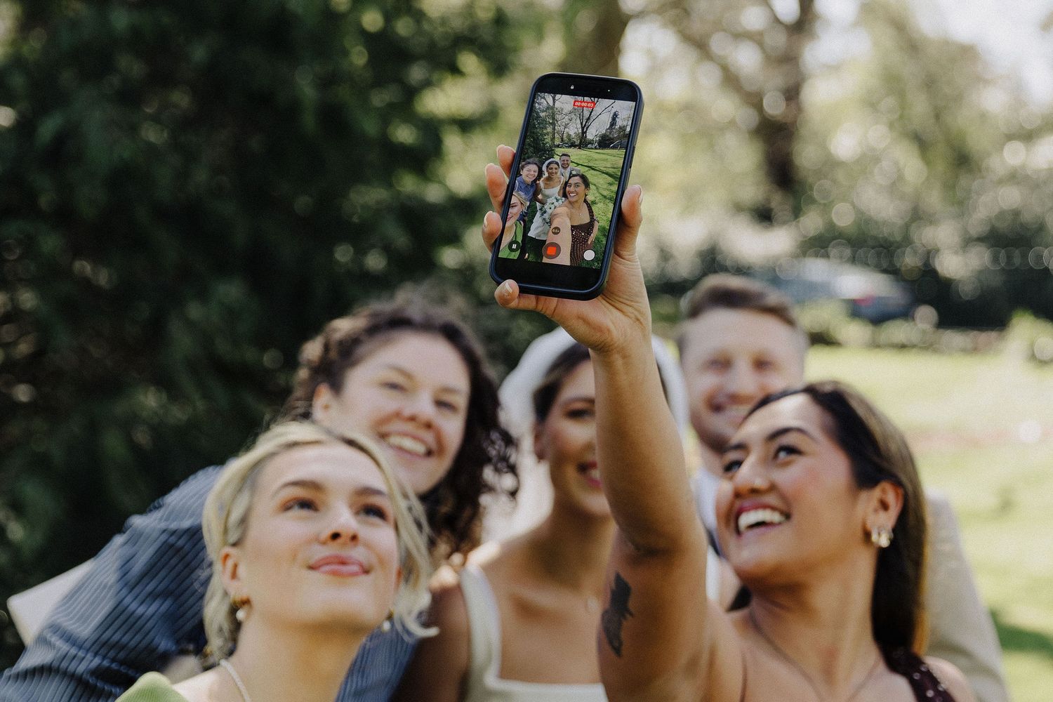 A group of smiling wedding guests taking a fun outdoor selfie together on a smartphone.