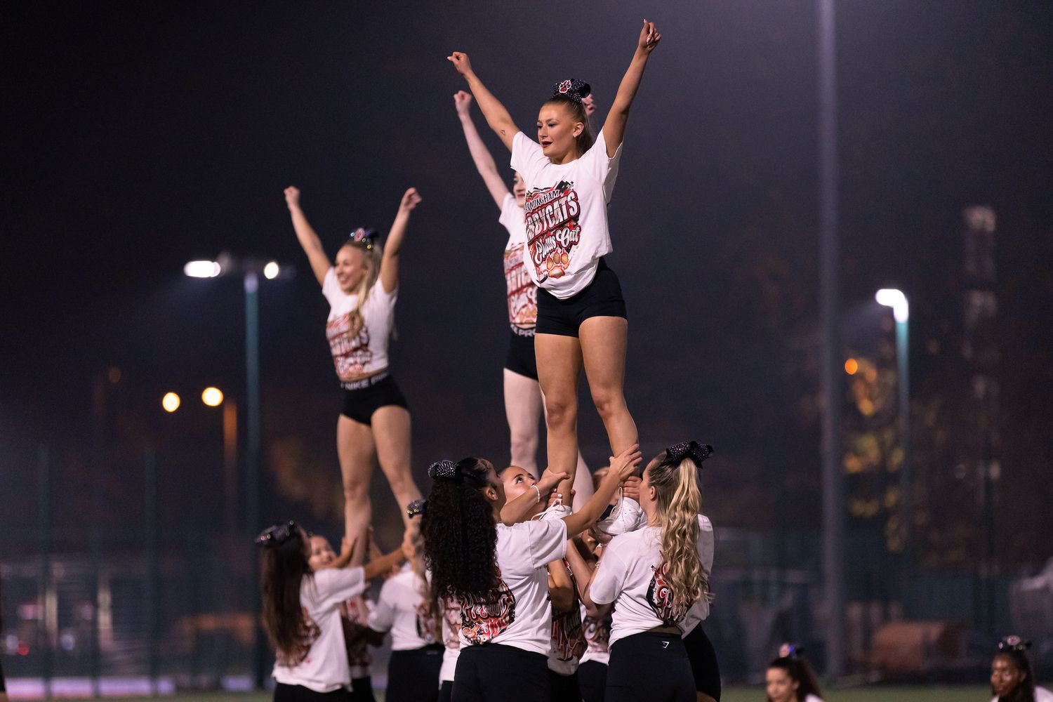 Cheerleaders perform synchronized stunts and lifts during a nighttime sporting event under bright stadium lights.