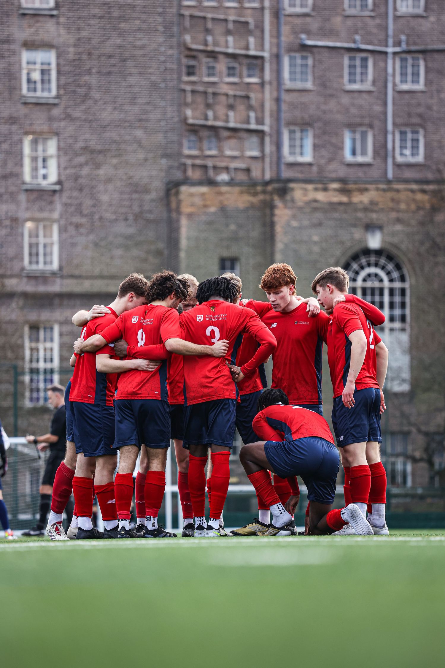 Soccer team huddles together in red uniforms in front of a historic brick building.