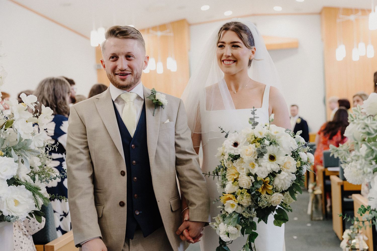 Smiling newlyweds walk down the aisle, groom in tan suit, bride holding white floral bouquet with veil.