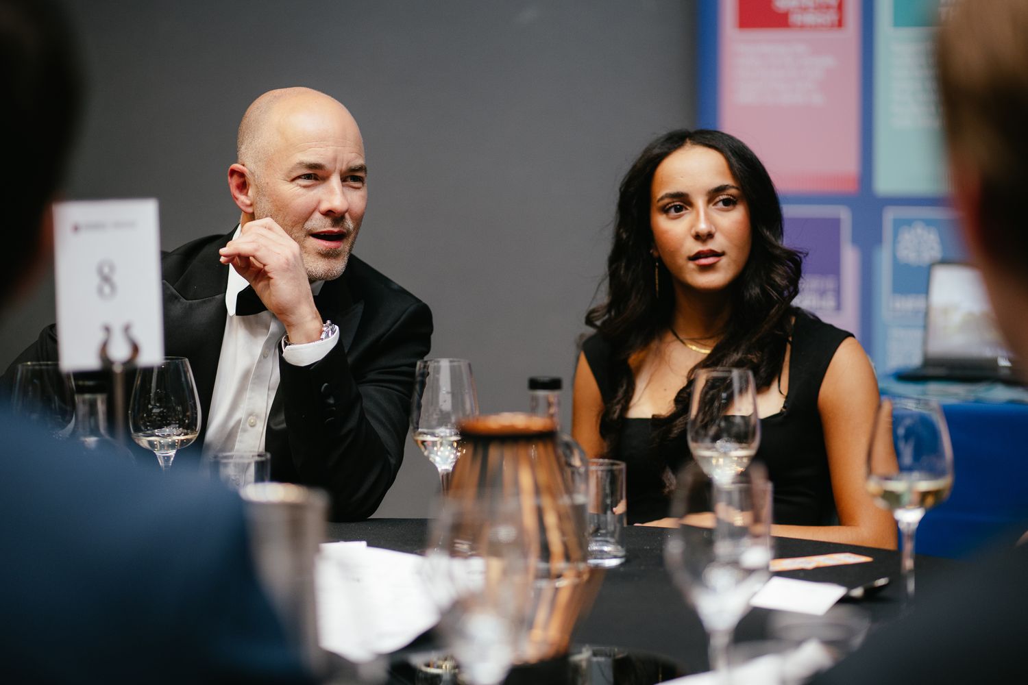 Two people sit at a dining table with wine glasses during what appears to be a business meeting or discussion.