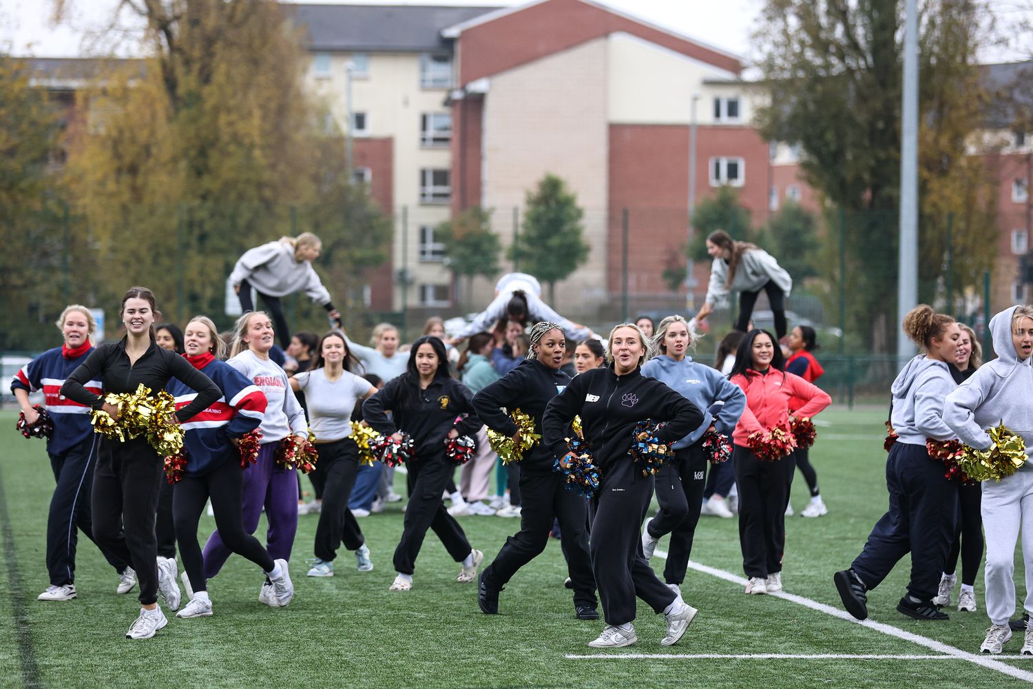 Cheerleaders practice their routine on a sports field during an autumn day with apartment buildings in the background.