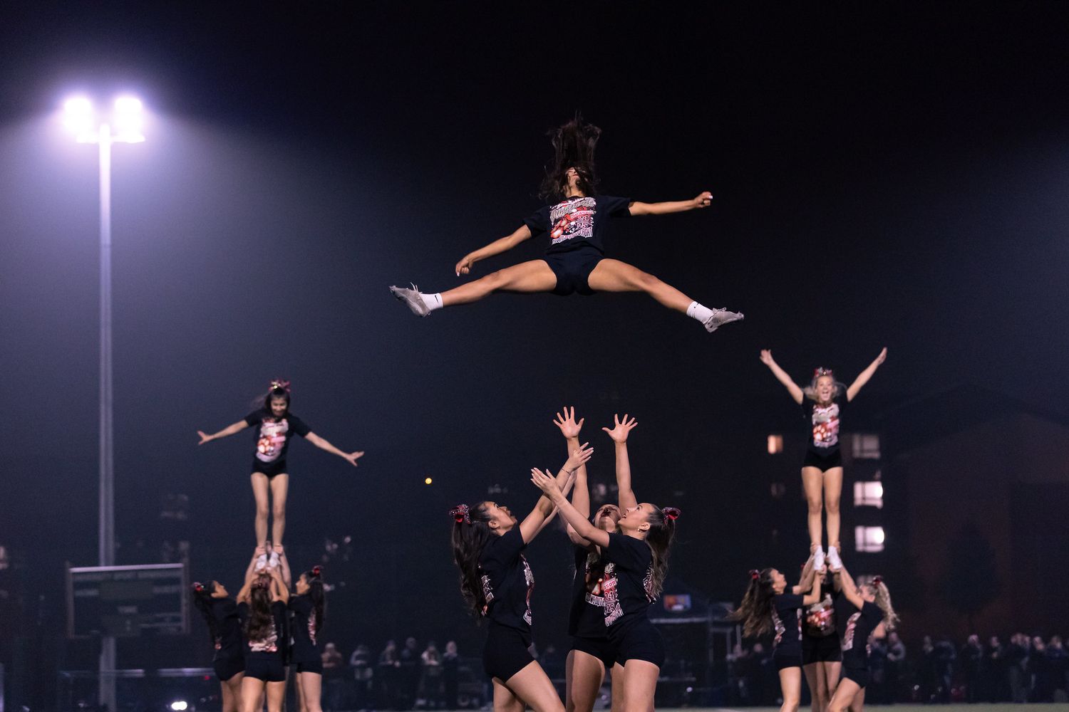 Cheerleaders executing complex aerial stunts during a nighttime performance under floodlights.