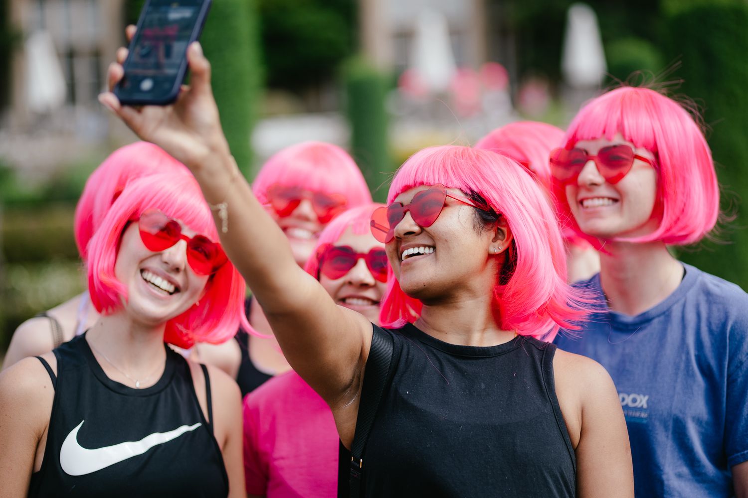 Group wearing matching pink wigs and heart sunglasses takes a selfie together.