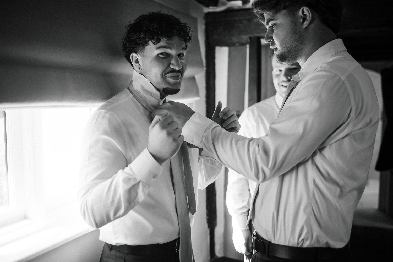 Two men in white dress shirts helping each other prepare for a wedding in black and white photo.