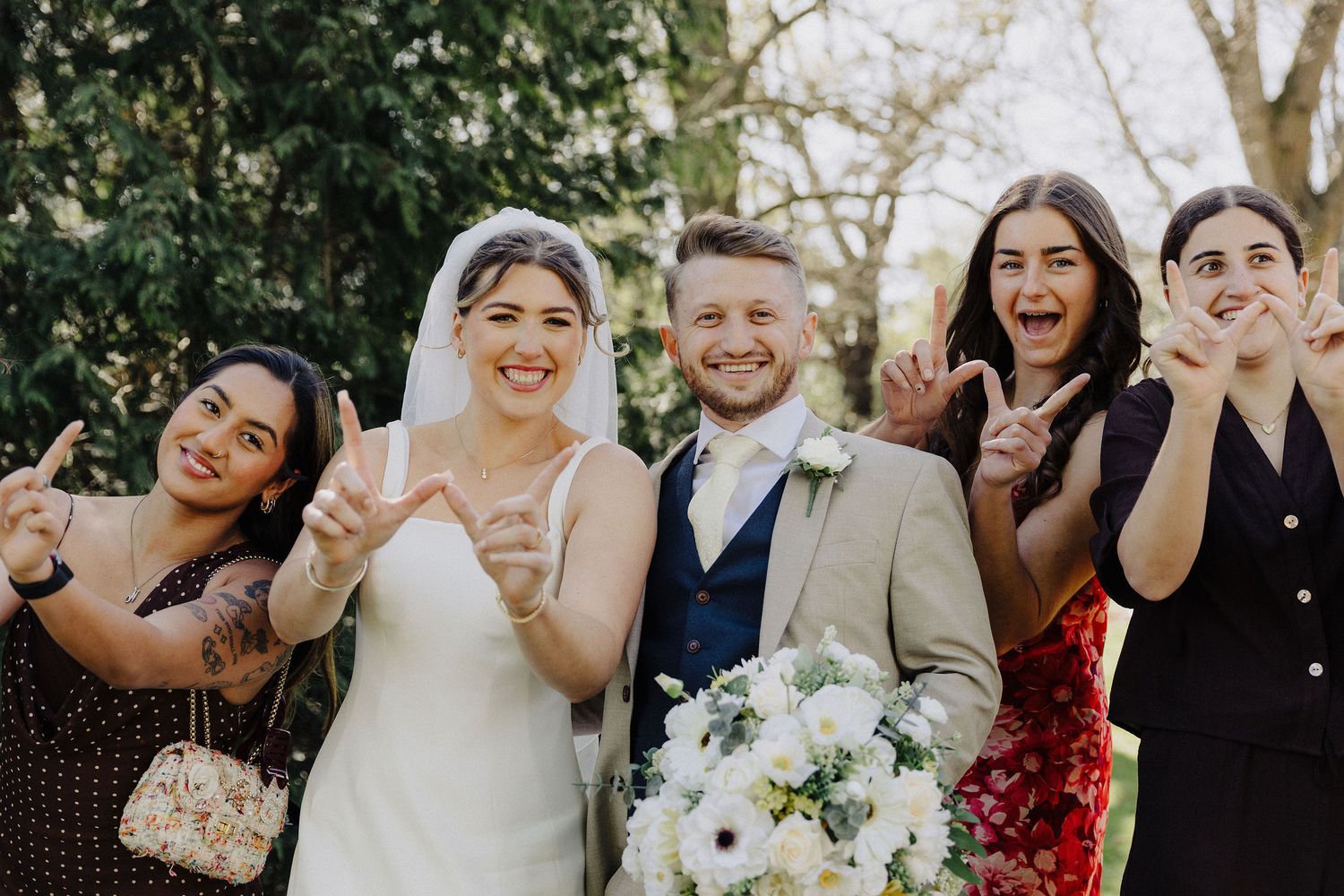 Smiling bride holding white bouquet poses with groom and three friends making peace signs outdoors.
