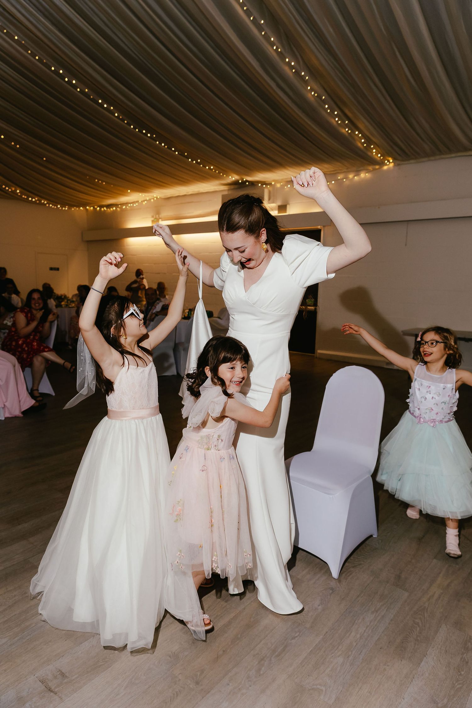 Bride dancing joyfully with flower girls in white and pink dresses at a wedding reception.