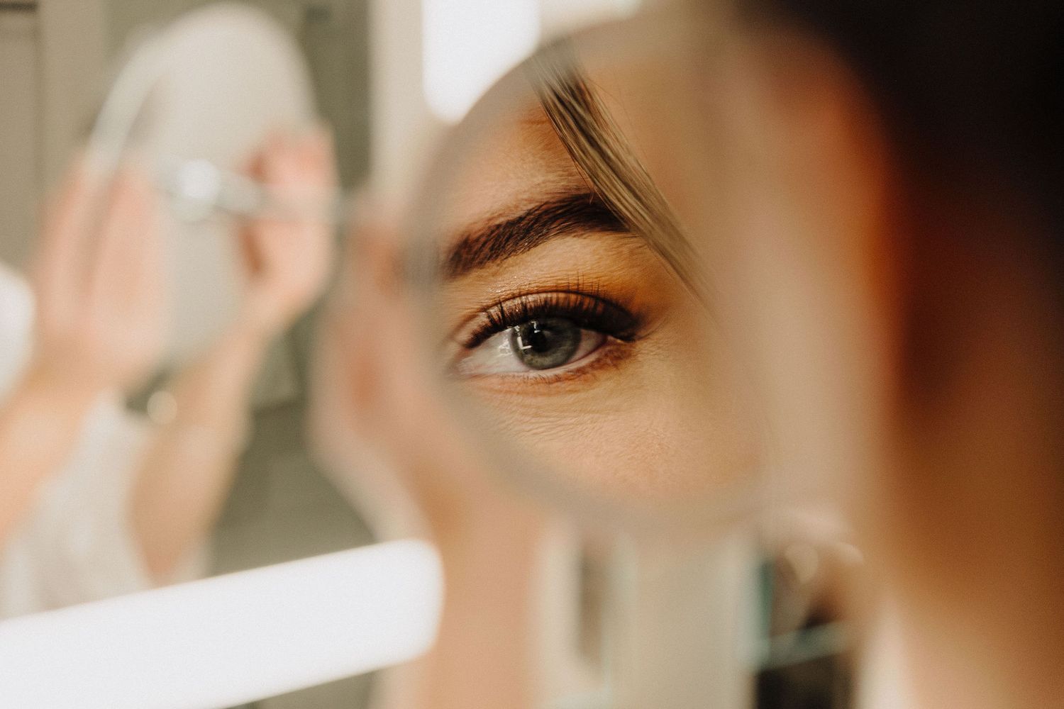 Close-up of a woman applying eye makeup while looking into a mirror, showcasing detailed eye with brown eyeshadow.