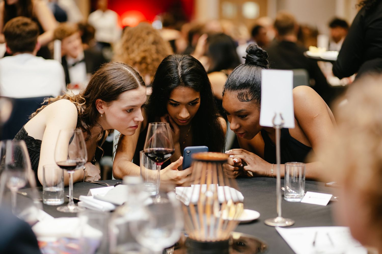 A group of friends huddle together looking at a phone screen during dinner at a restaurant table with wine glasses.