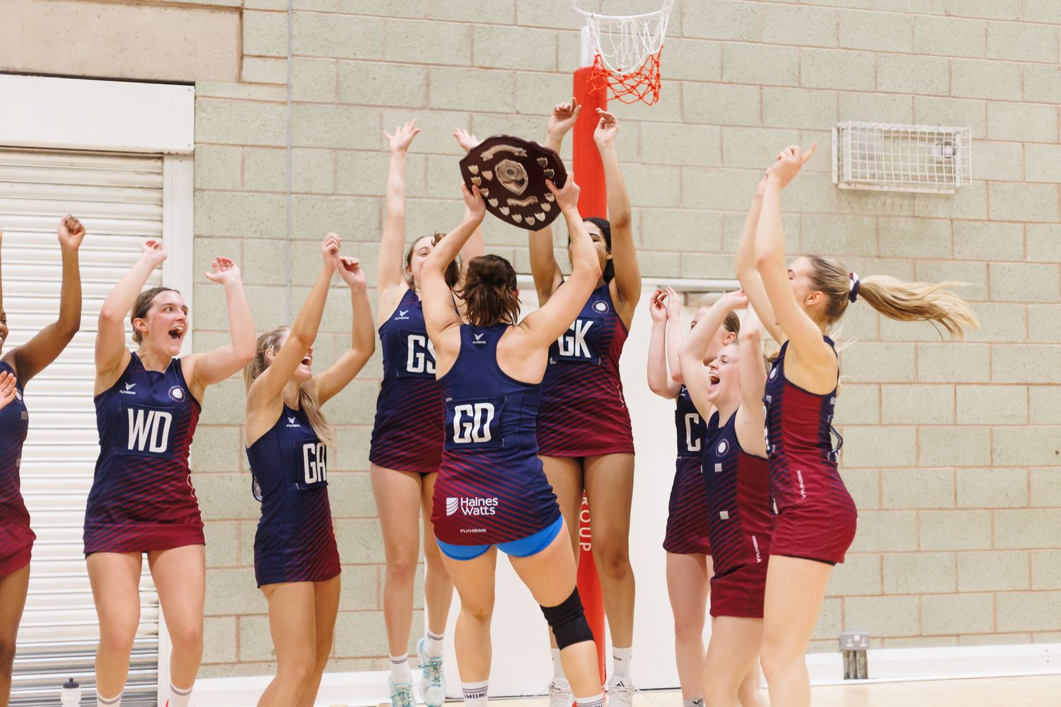 Netball players practice jumping drills in an indoor sports facility.
