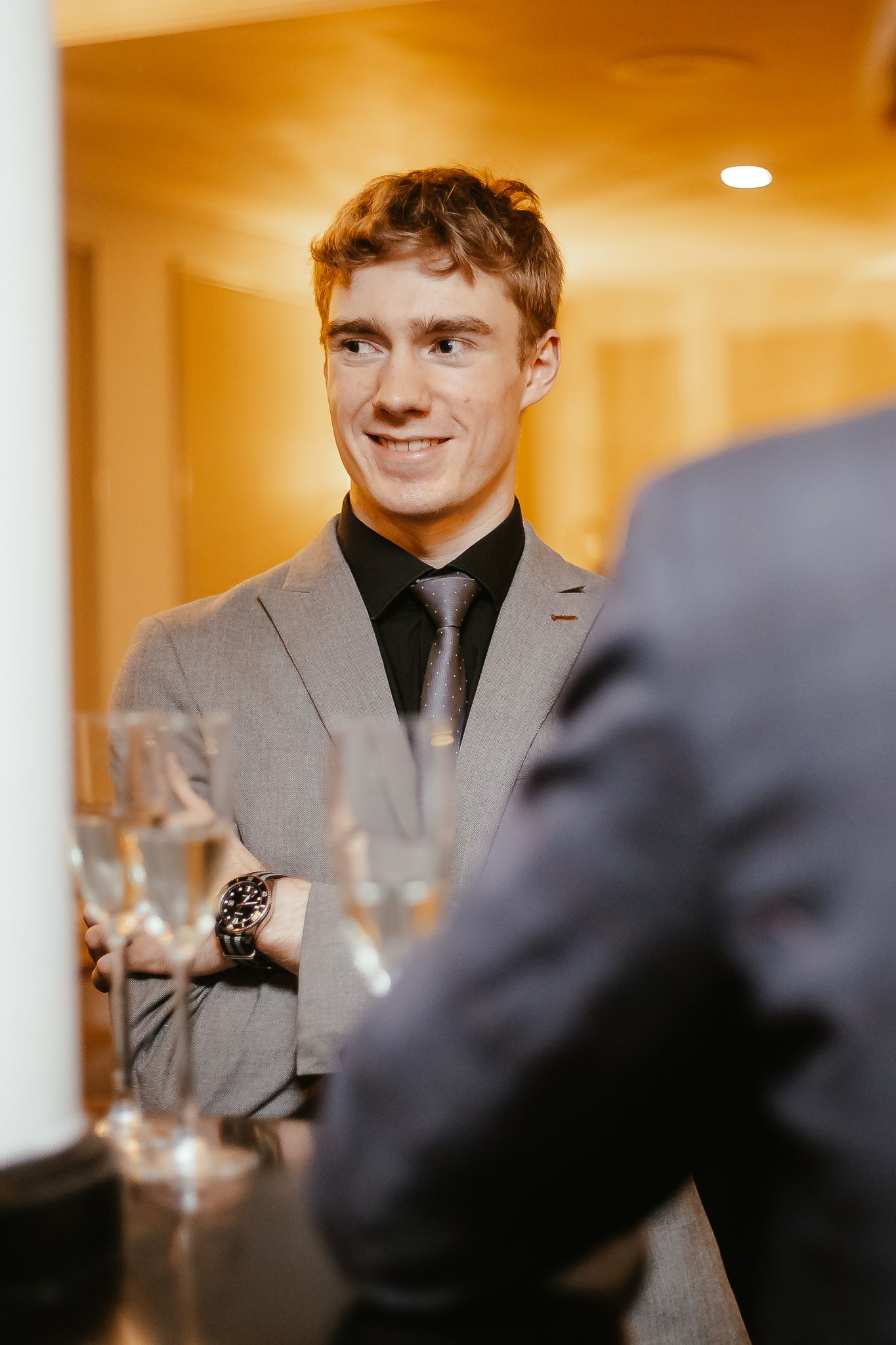 Guest in grey suit smiling at formal event with wine glasses on table.