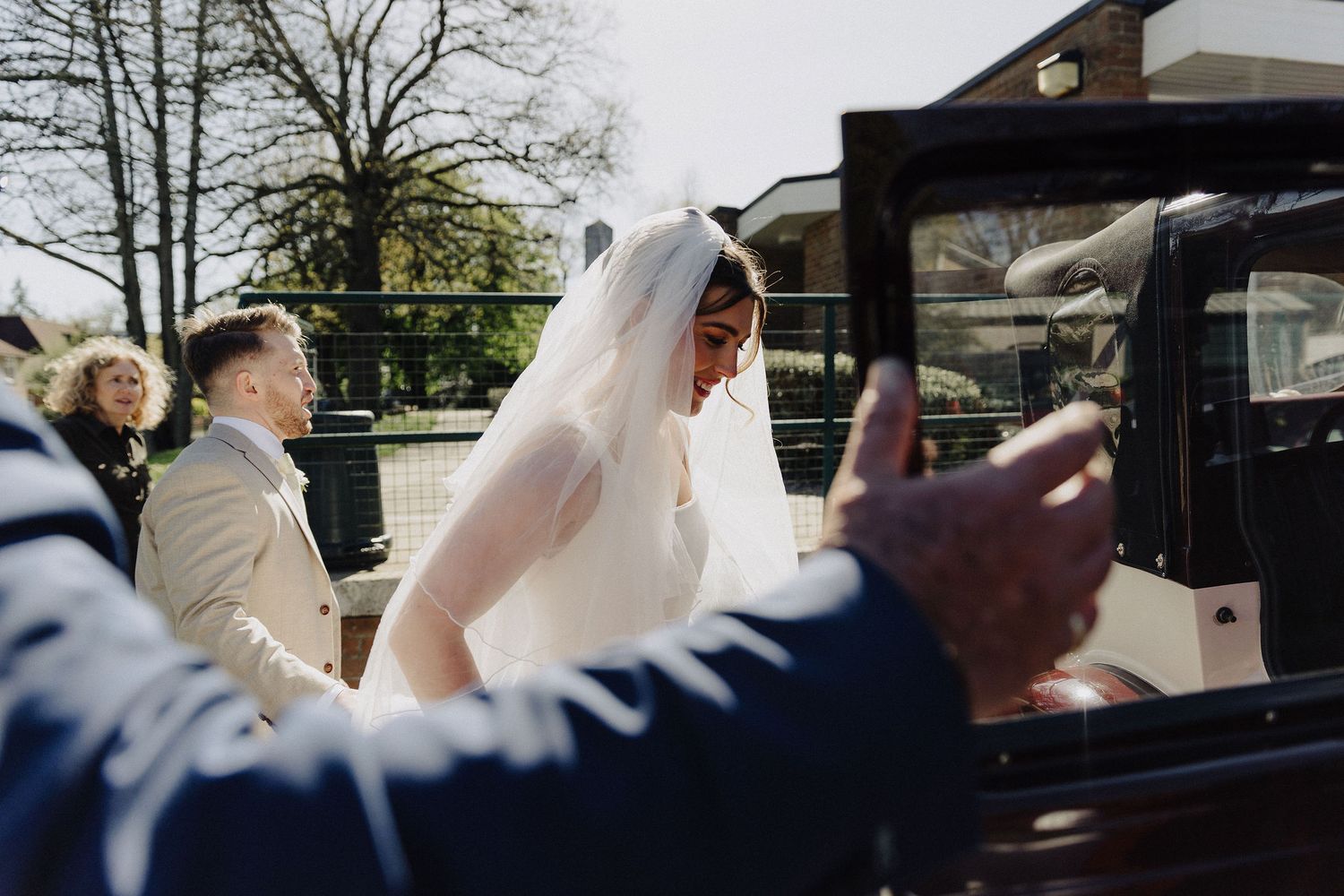 Bride in white gown and veil arriving at church in a vintage black wedding car on a sunny day.