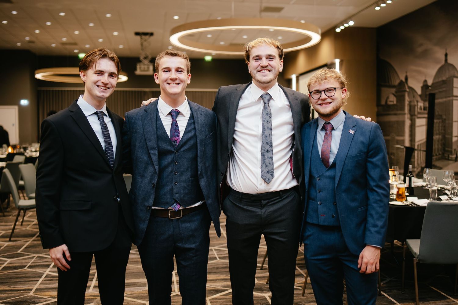 A group of groomsmen in dark suits and ties pose together at an elegant indoor venue.