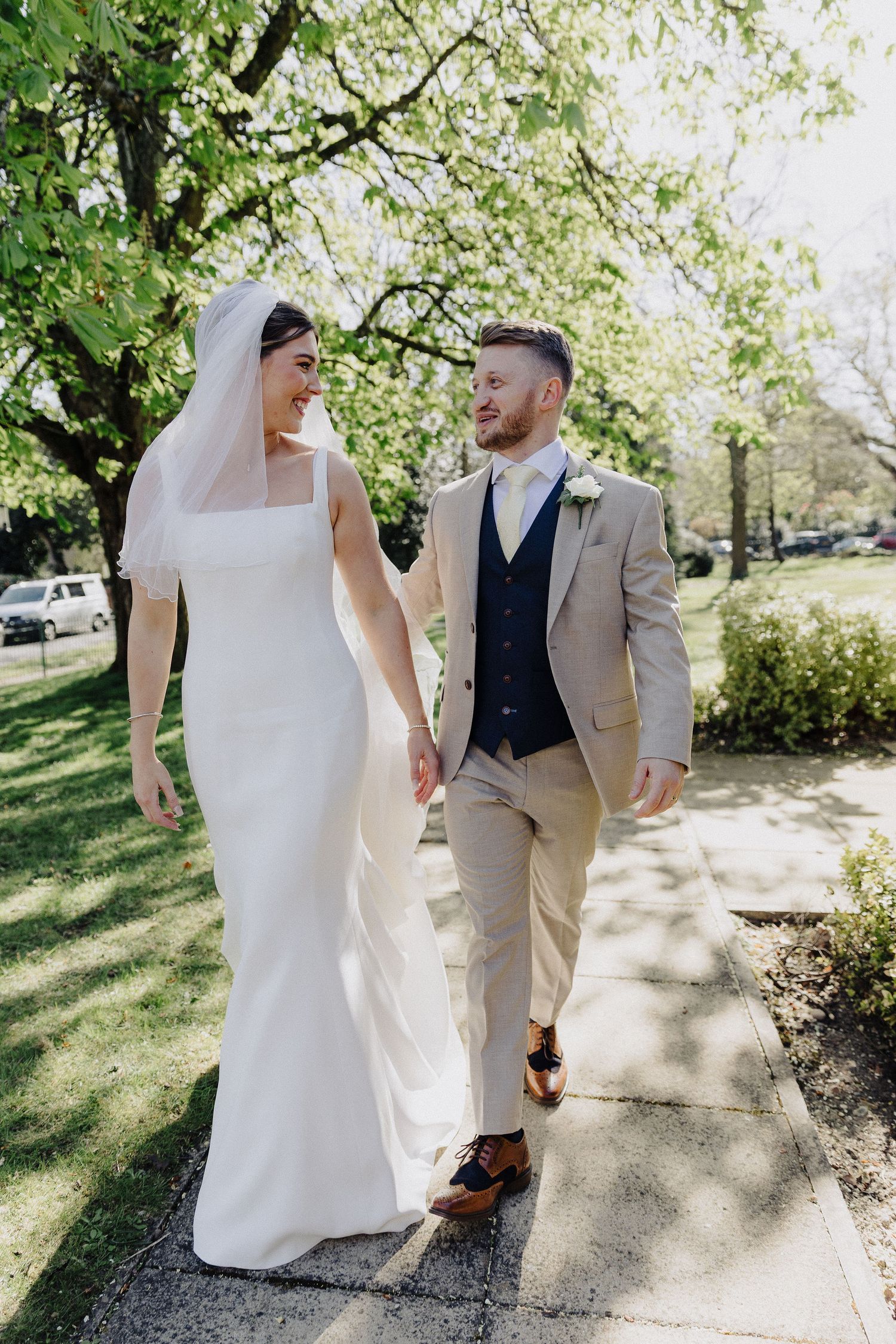 Bride in white gown and veil walks hand-in-hand with groom in tan suit on sunny tree-lined sidewalk.