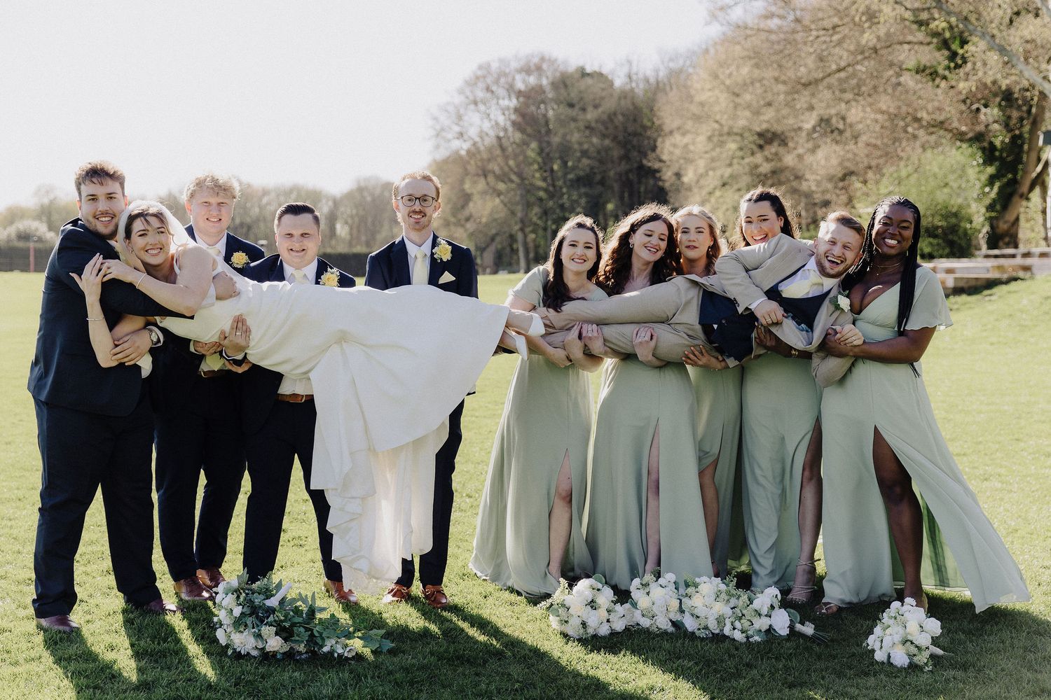 Wedding party posing outdoors, groomsmen lifting bride while bridesmaids in sage green dresses celebrate on green lawn.