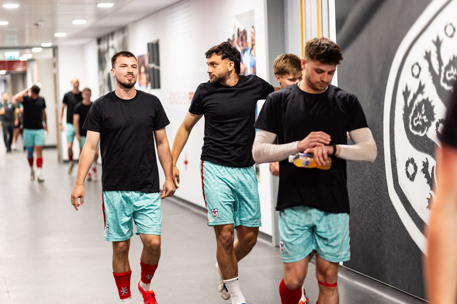 Athletes walking through modern sports facility hallway wearing teal and black uniforms.