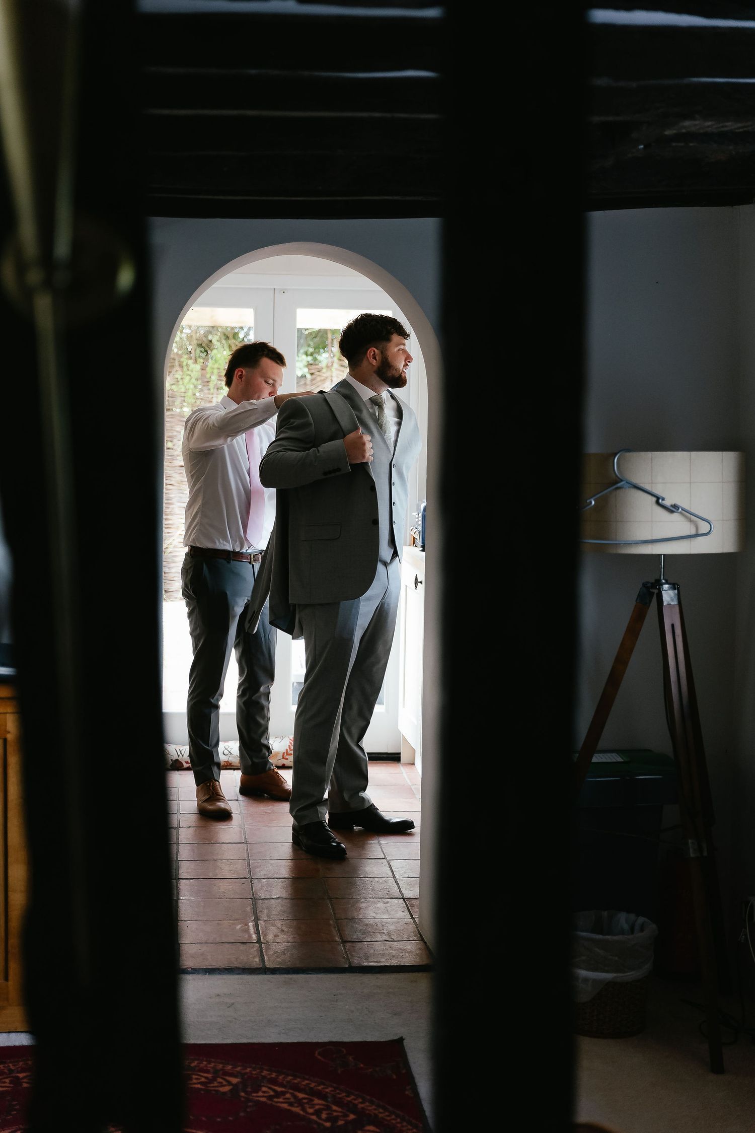 A groom in a gray suit gets help from a groomsman adjusting his jacket in a rustic venue doorway.