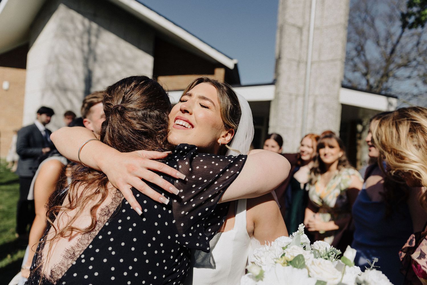 Smiling bride in white dress shares heartfelt hug with guest while holding white bouquet after ceremony.