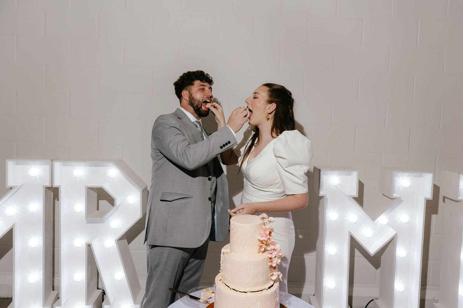 Bride and groom feeding each other wedding cake between illuminated MR and MRS letters.