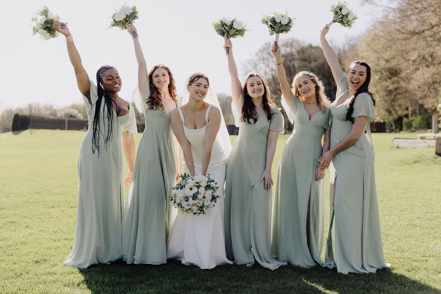 Bride and four bridesmaids in sage green dresses celebrating outdoors, raising bouquets of white flowers on a sunny day.