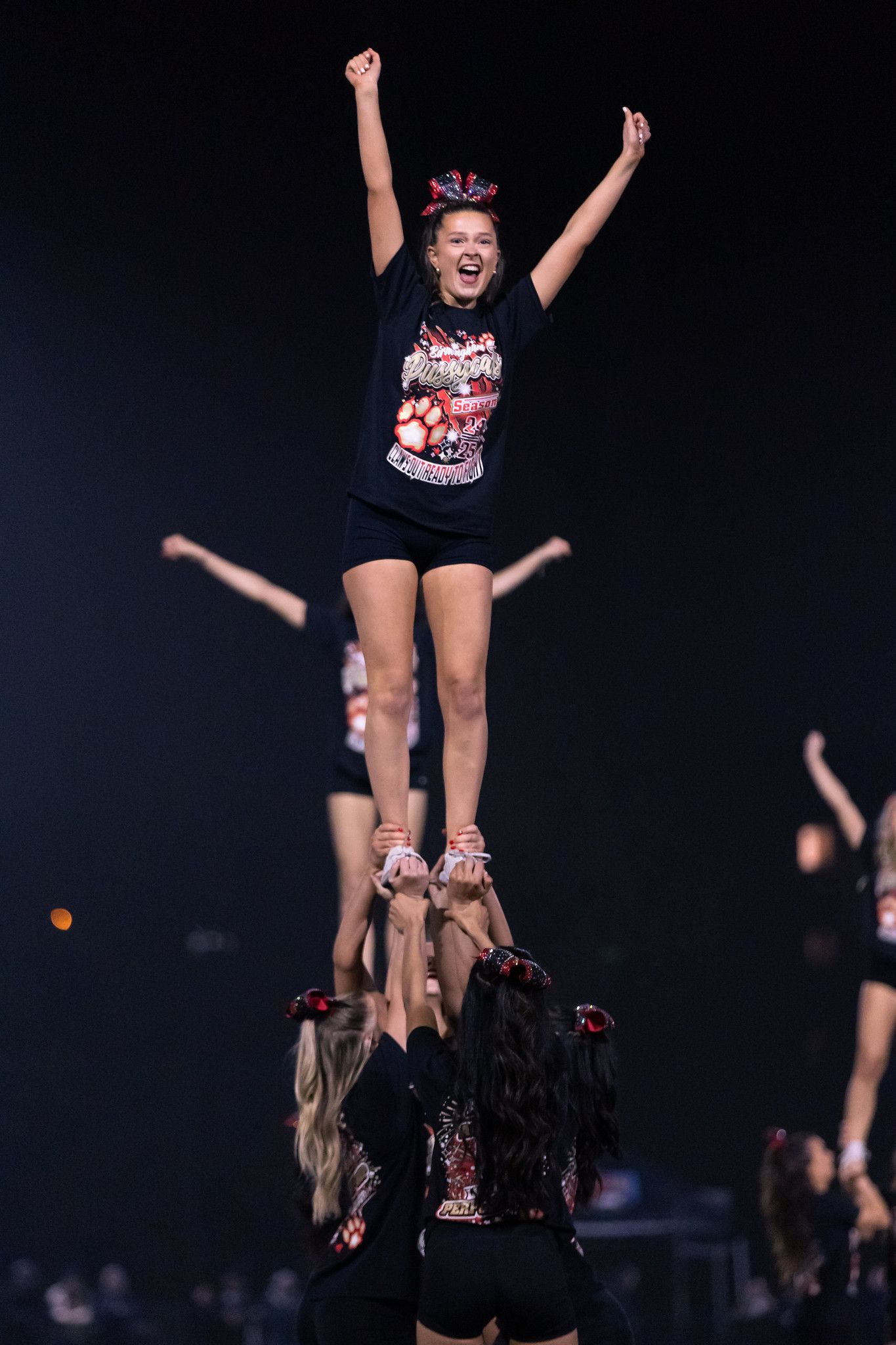 Cheerleaders perform synchronized stunts and lifts during a high-energy performance on a dark stage.