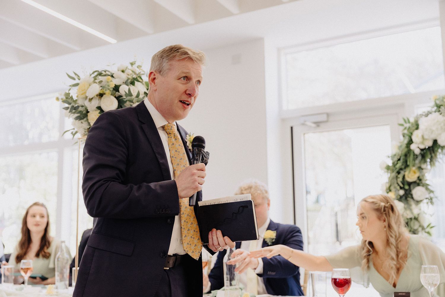 Man in navy suit giving a speech at a wedding reception while holding a microphone and papers.