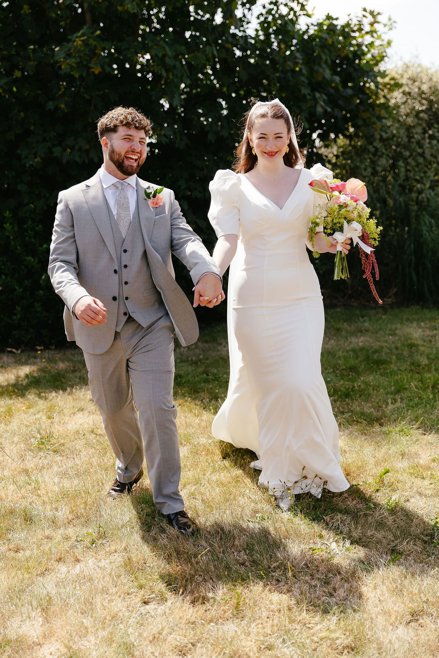 A happy bride and groom walk hand in hand outdoors on their sunny wedding day.