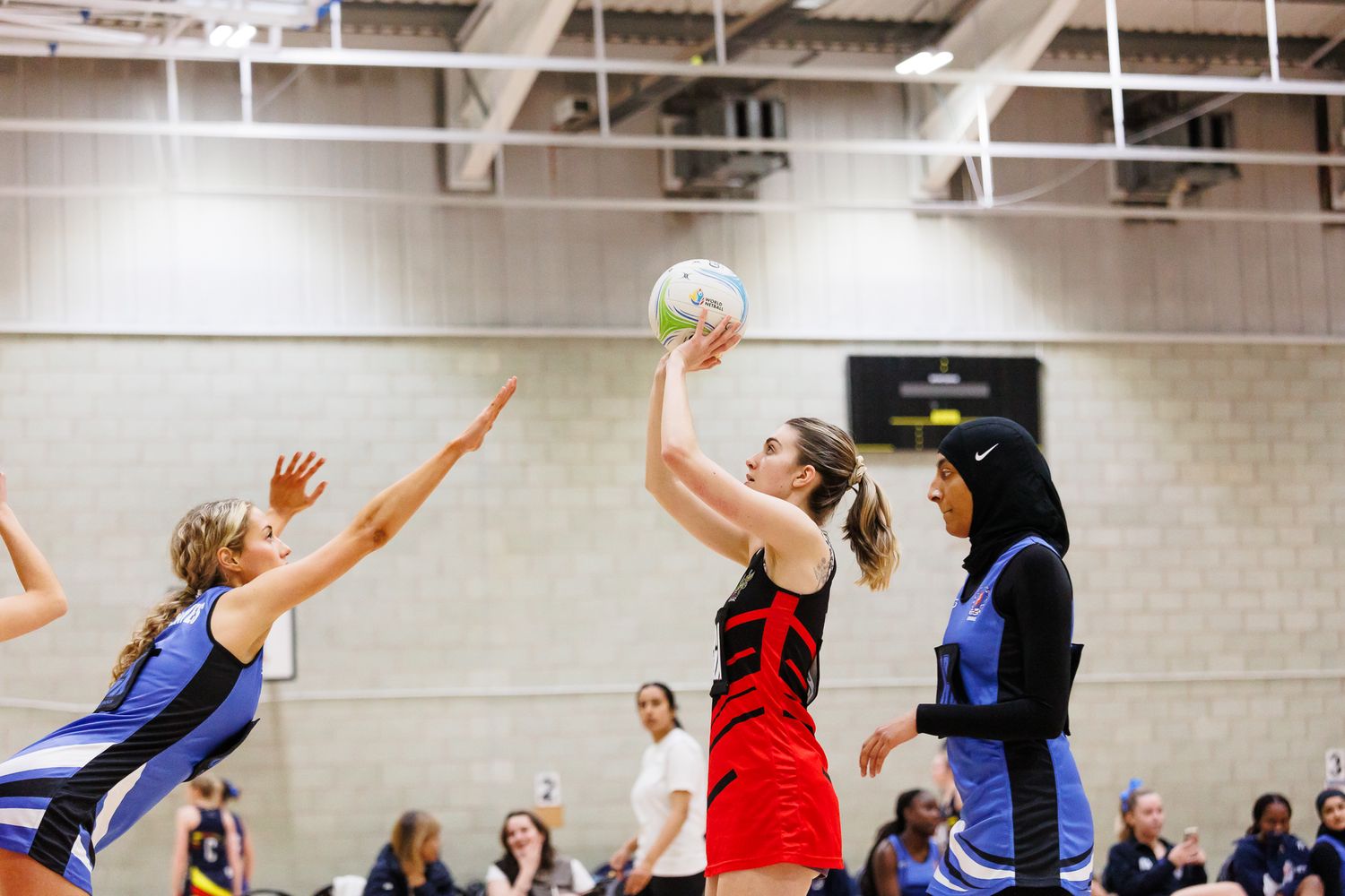 Athletes jumping and reaching for netball during an indoor sports match.