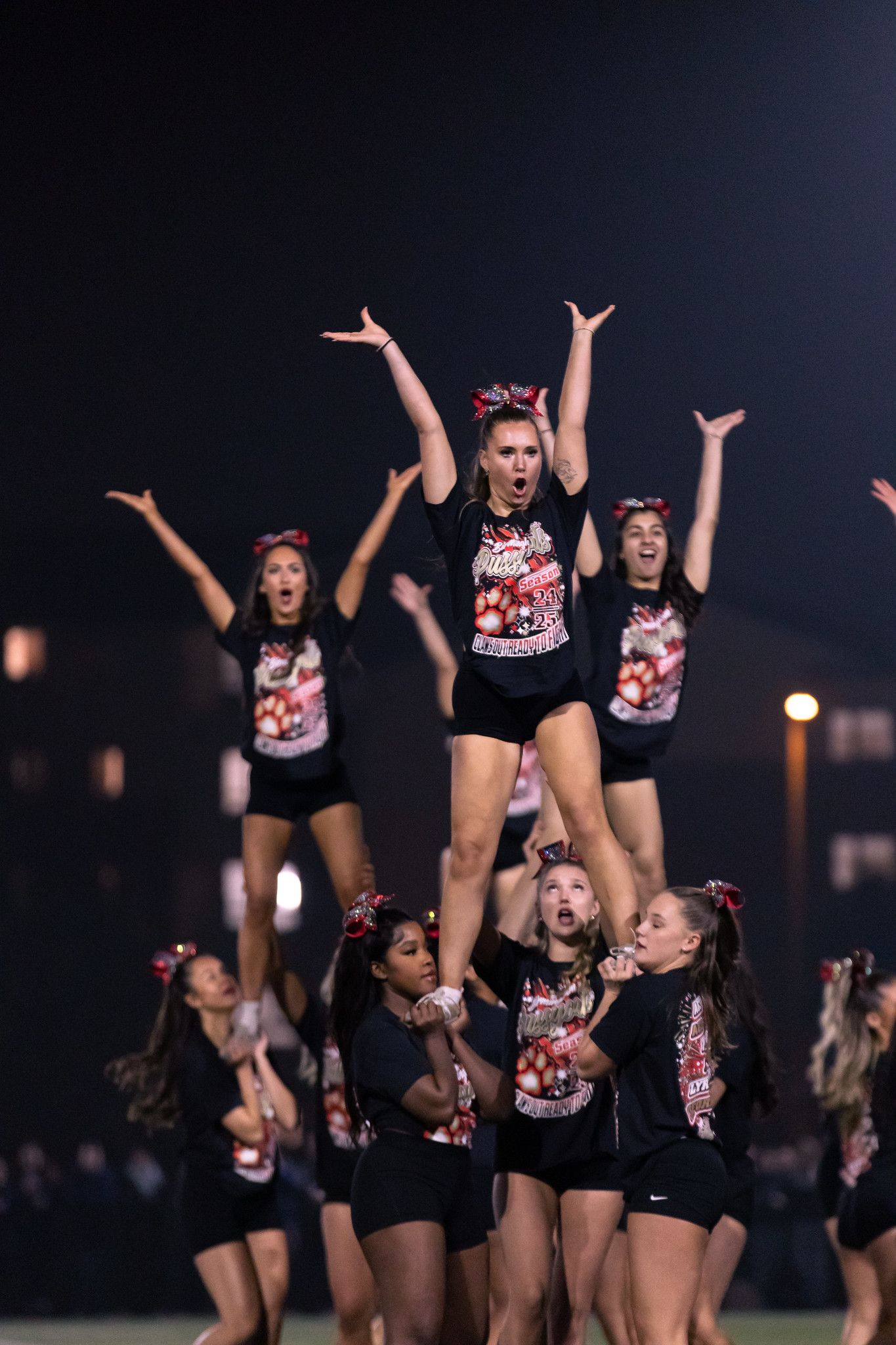 Dance team performs synchronized routine in black uniforms with matching pink and white tops during nighttime show.
