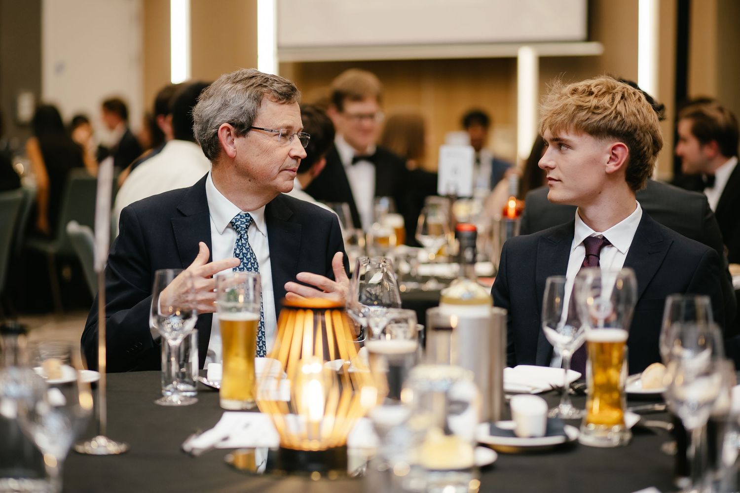 Business professionals gathered around tables at a formal dinner event in a well-lit conference space.
