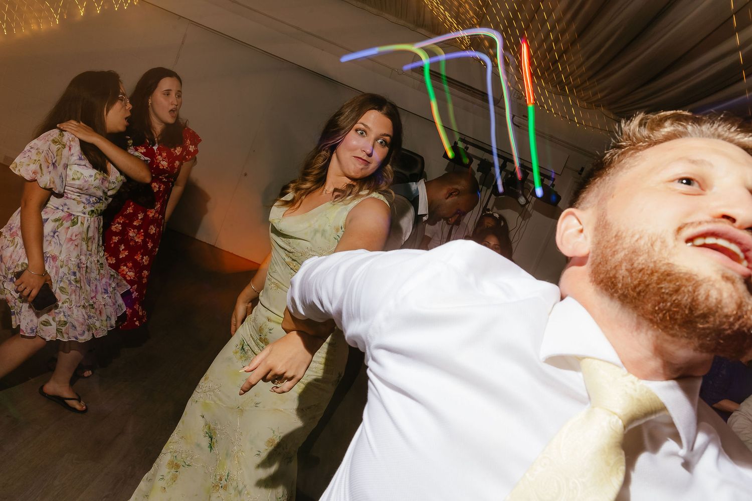 Guests dance energetically at a wedding reception with colorful glow sticks and festive lighting in background.