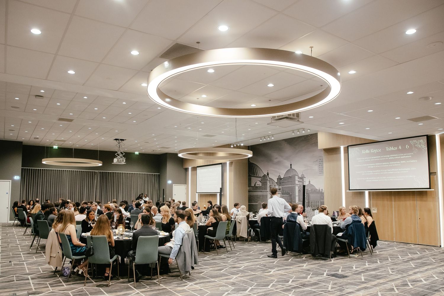Professional conference room with multiple round tables and chairs arranged in rows under modern recessed lighting.