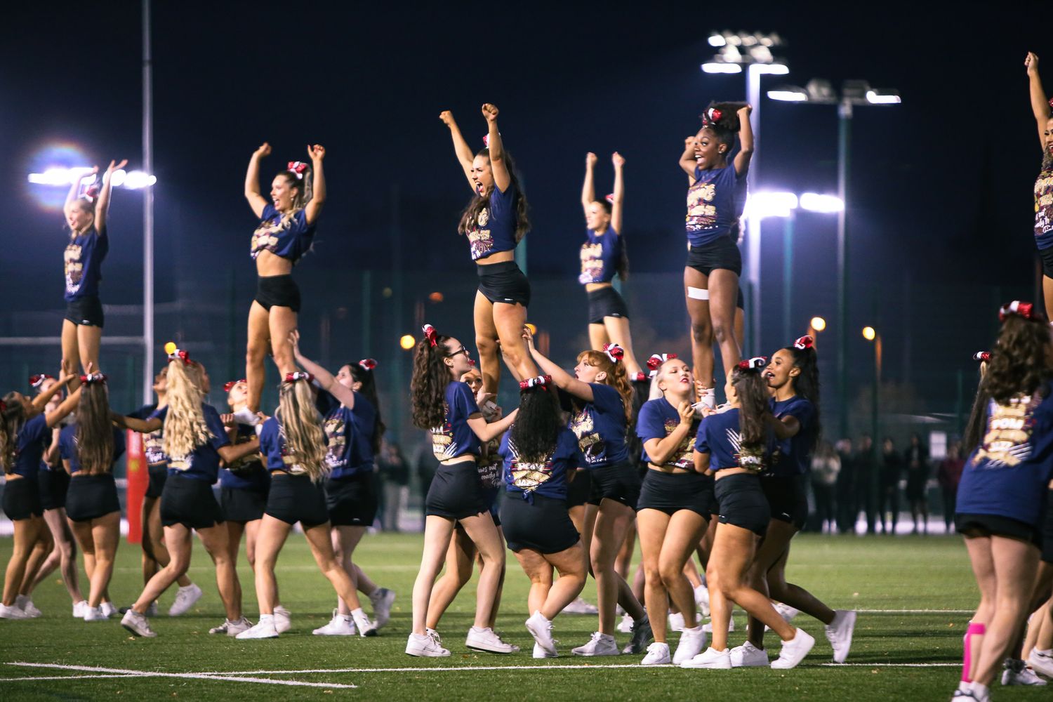 Cheerleaders perform synchronized jumps and dance moves during a nighttime performance on an illuminated field.