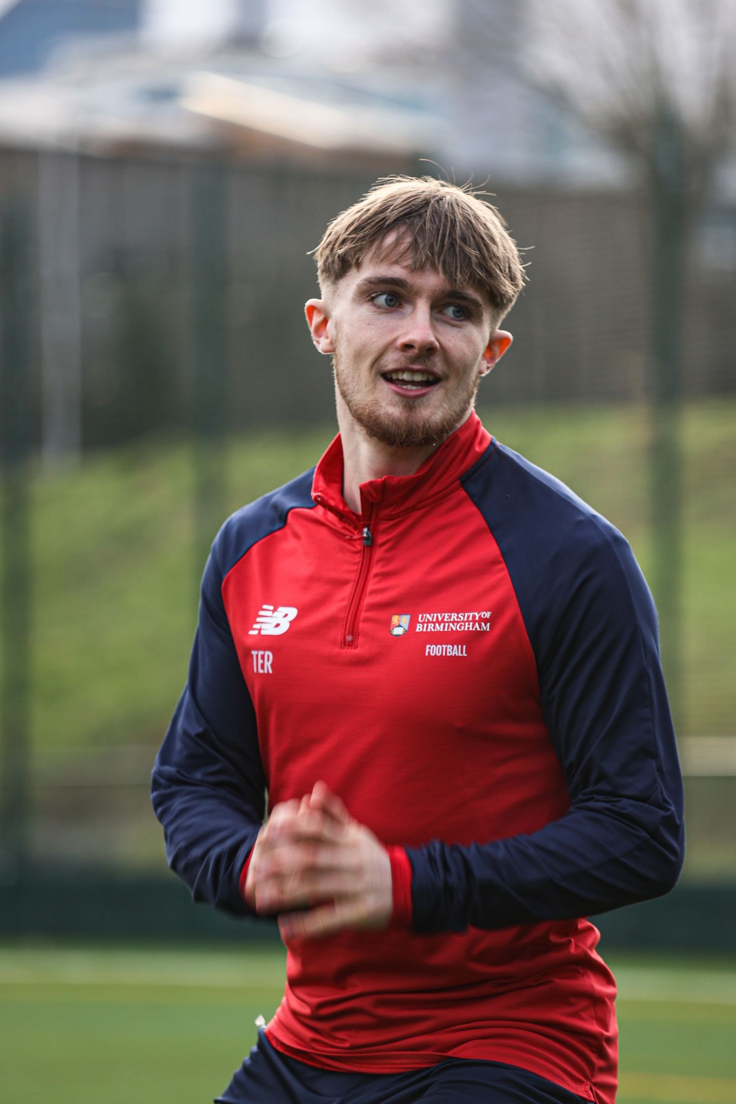 Athlete in red and navy New Balance training gear running outdoors on a sports field.