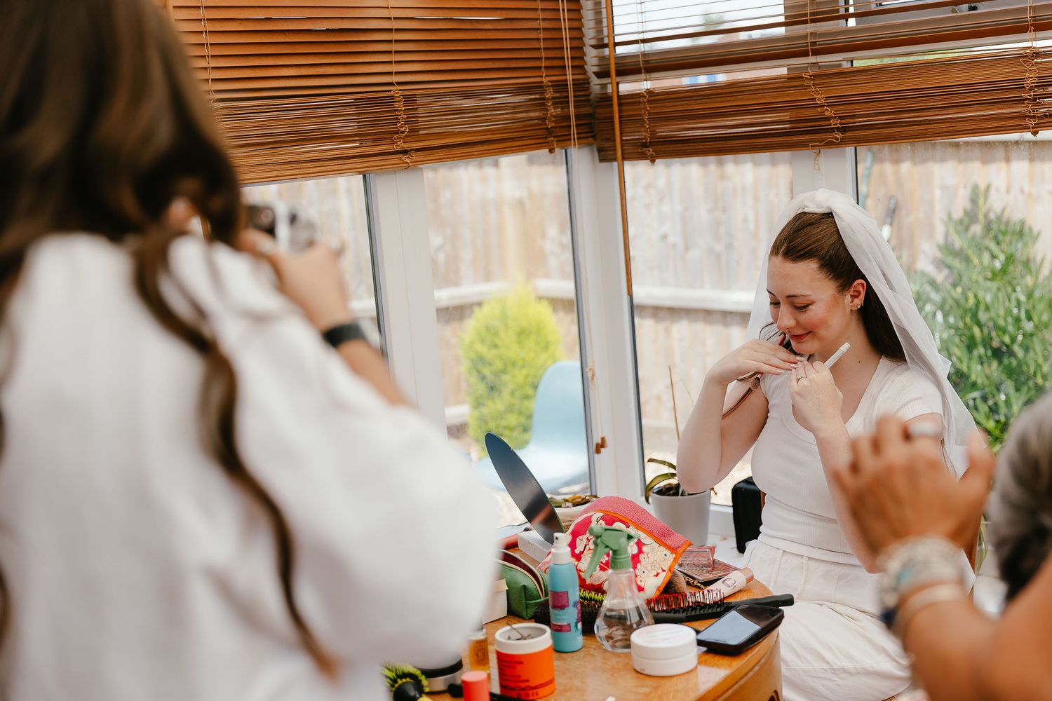 Bride in white dress laughing while getting ready at a table with makeup and beauty products before her wedding.