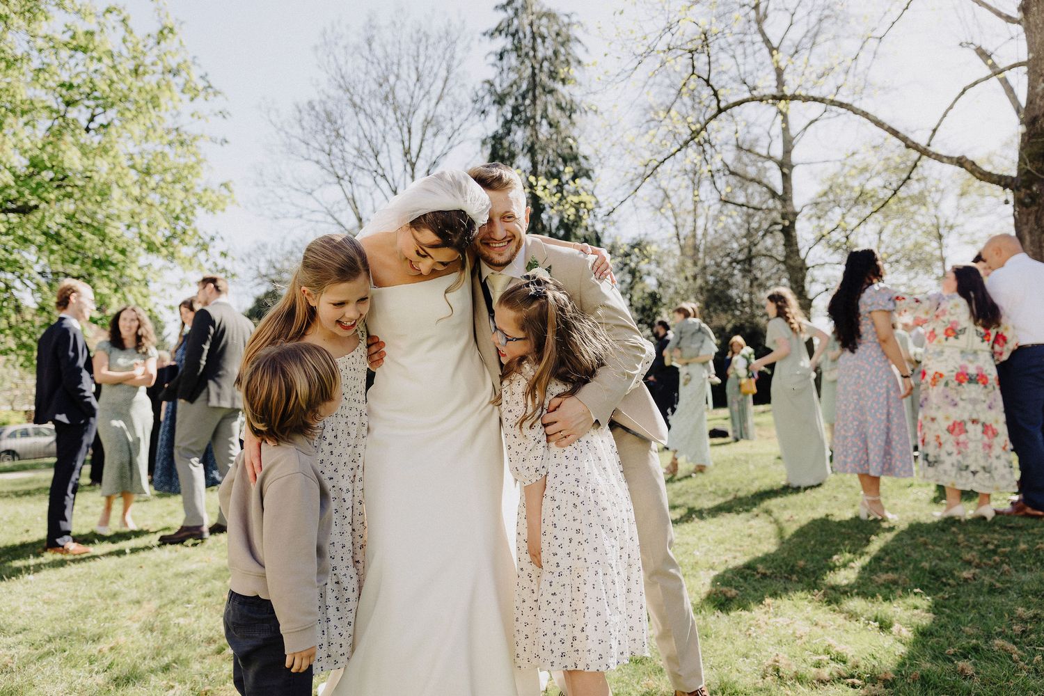 Bride and groom hugging children outdoors at wedding reception on sunny day with guests in background.
