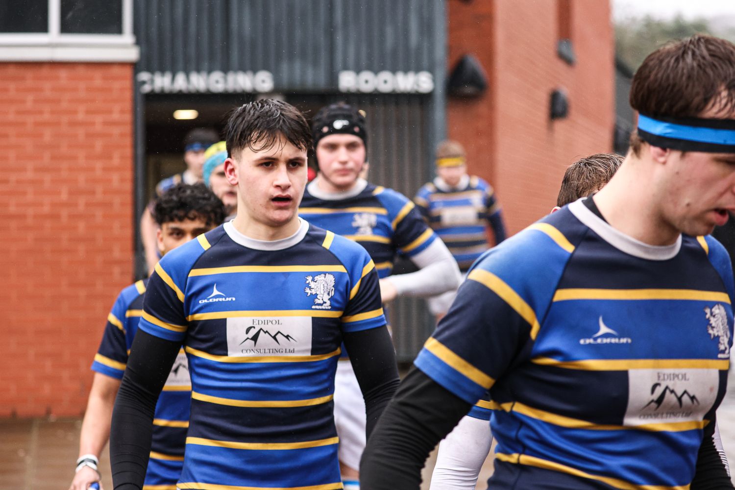 Rugby players line up for a play against a brick building backdrop.