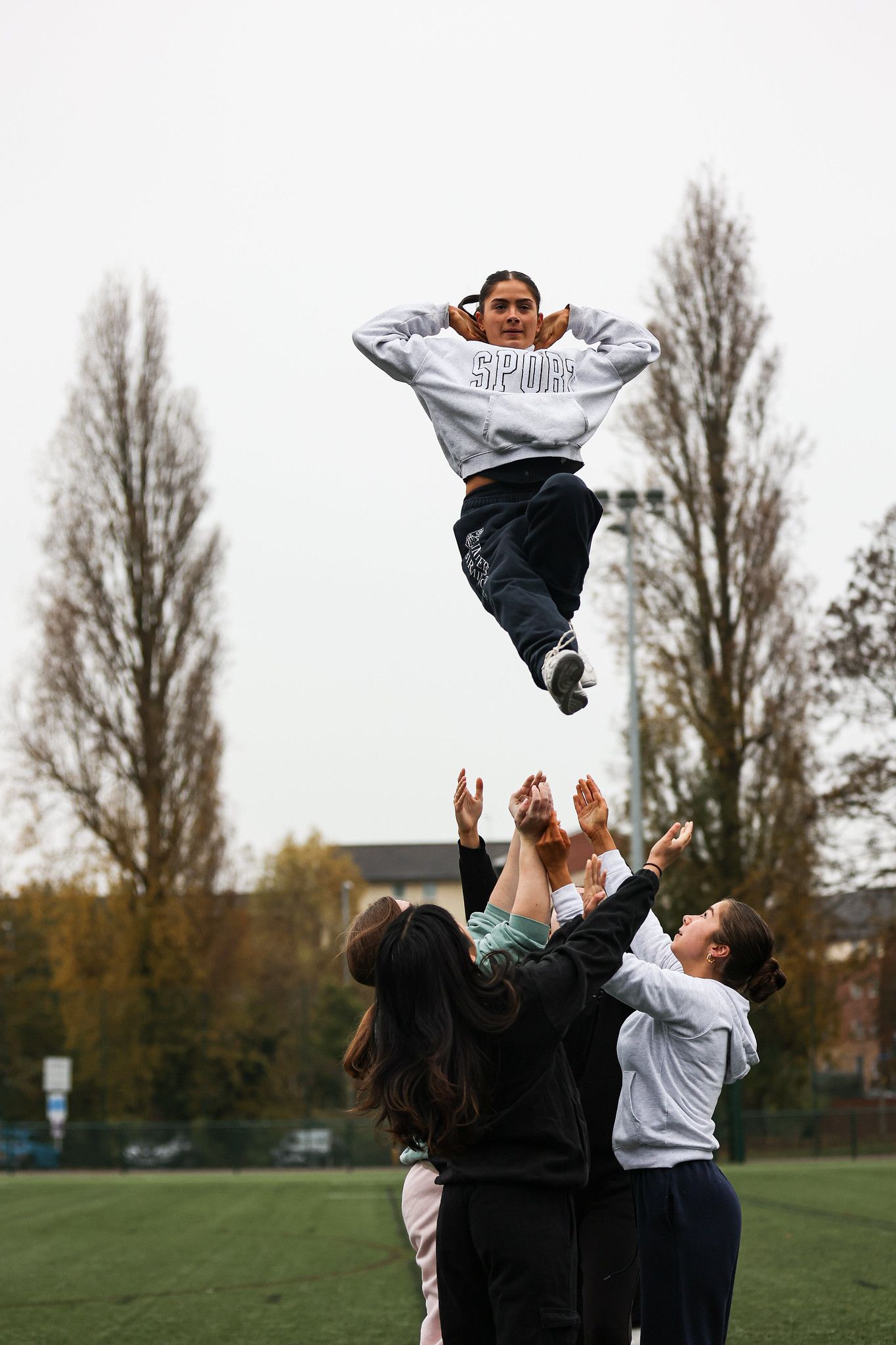 A group performs a cheerleading stunt on a grassy field, tossing someone high into the air against a backdrop of trees.