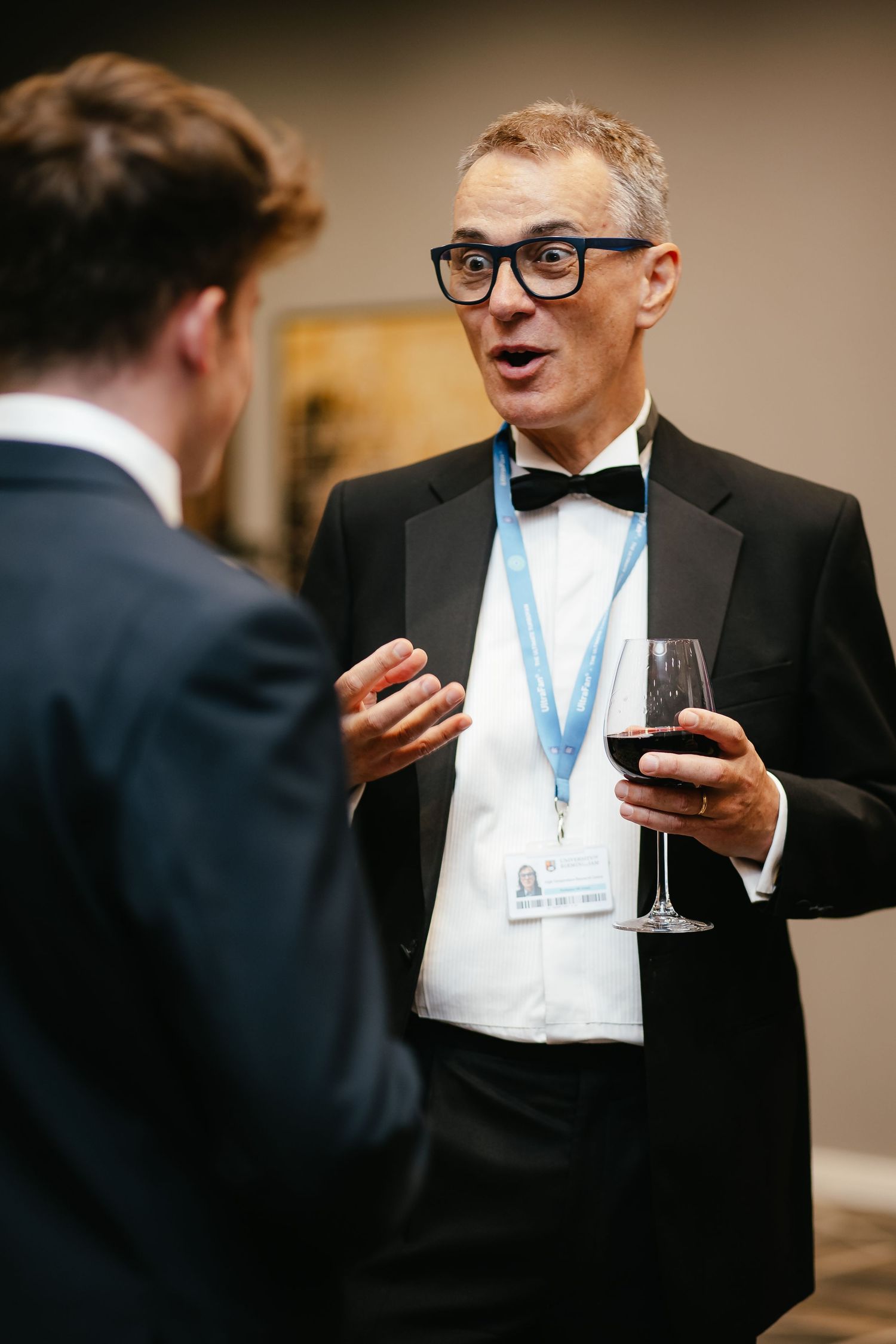 Well-dressed attendee with glasses engaged in conversation while holding wine at formal event.