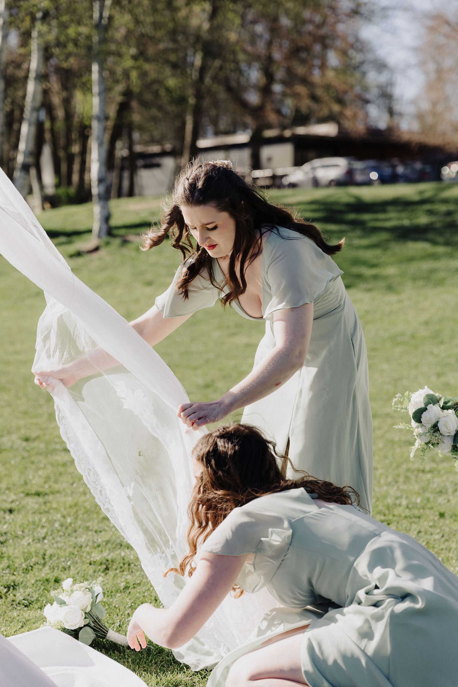 Two bridesmaids in sage green dresses arranging a white wedding gown on a sunny green lawn outdoors.