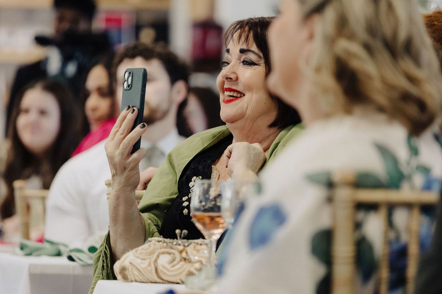 Woman smiling and taking a selfie with her phone at an elegant indoor event or celebration.