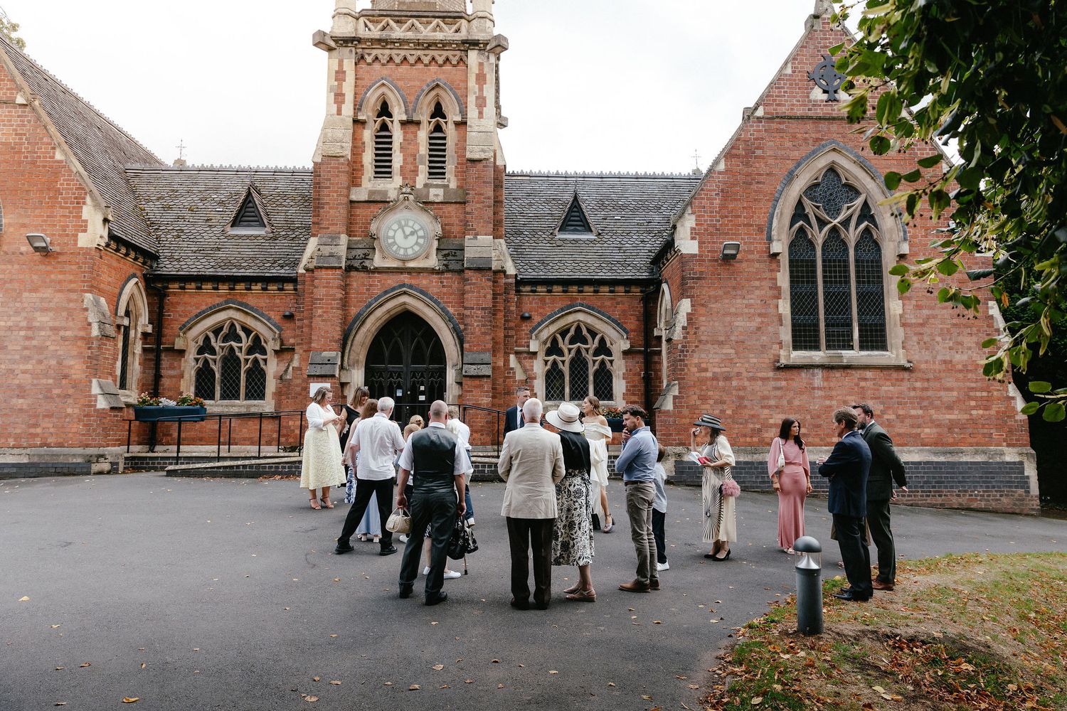 Wedding guests gather outside a historic red brick Gothic church with clock tower on a cloudy day.