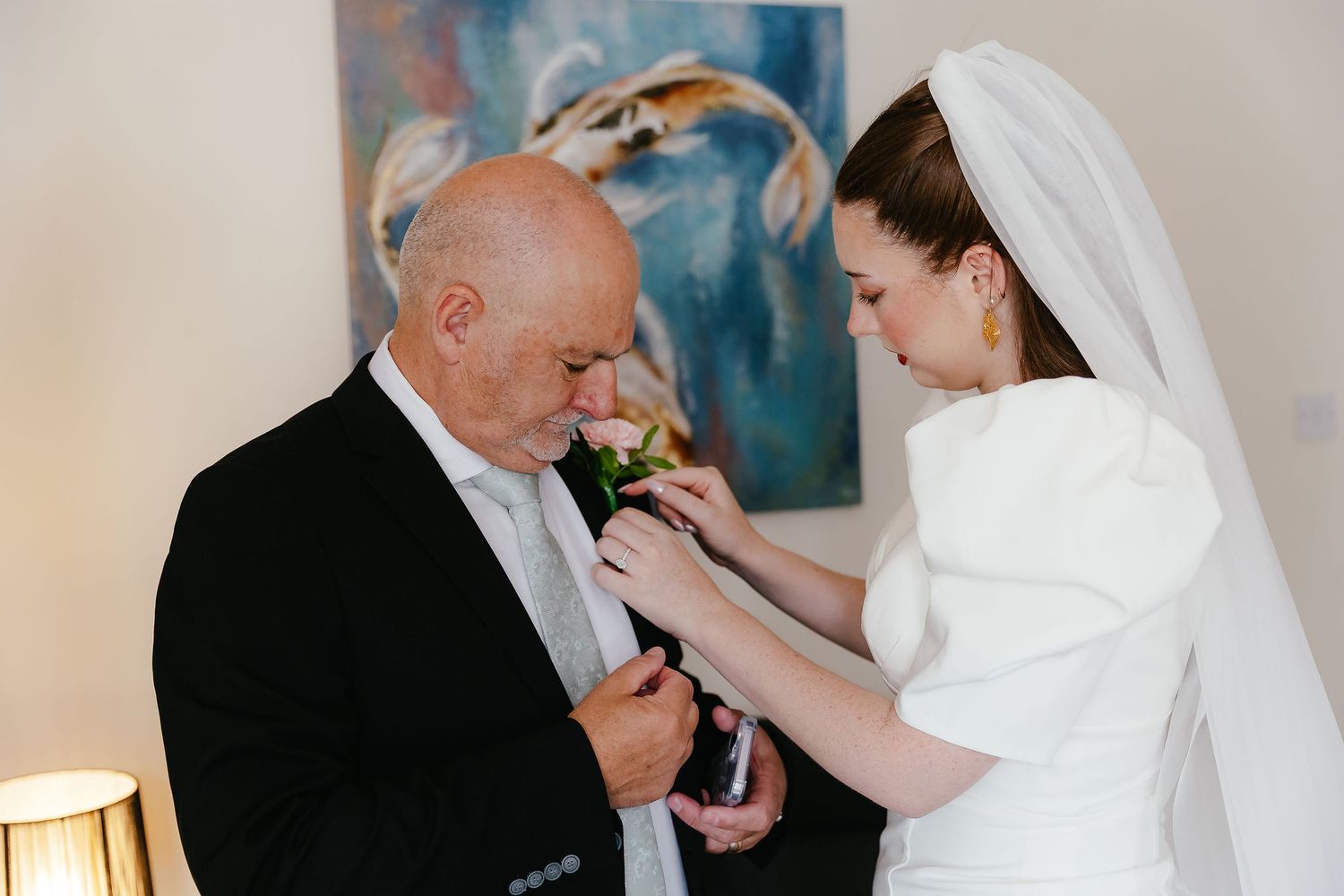 Bride in white gown pins boutonniere on older man in black suit before wedding ceremony.