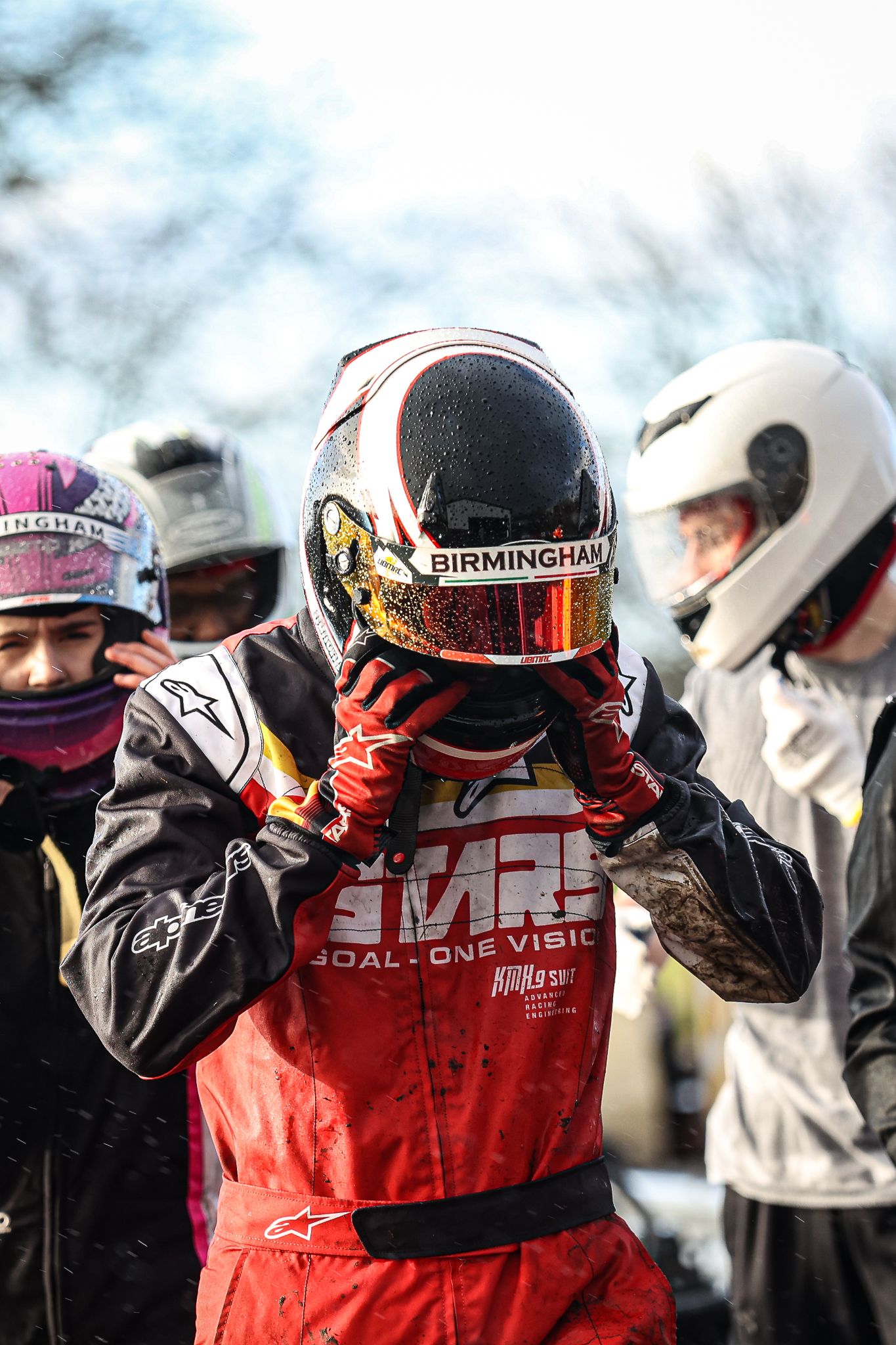 Group of racing drivers in helmets and racing suits gathered together trackside.