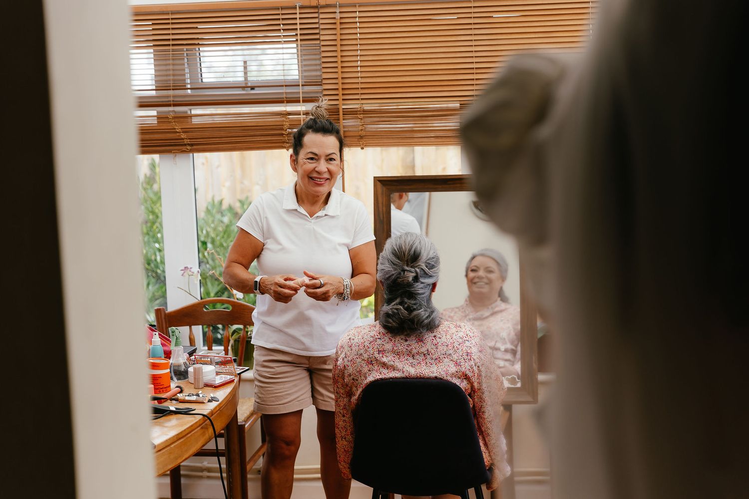 Woman in white top smiles at seated person getting hair done in a bright salon room with wooden blinds.