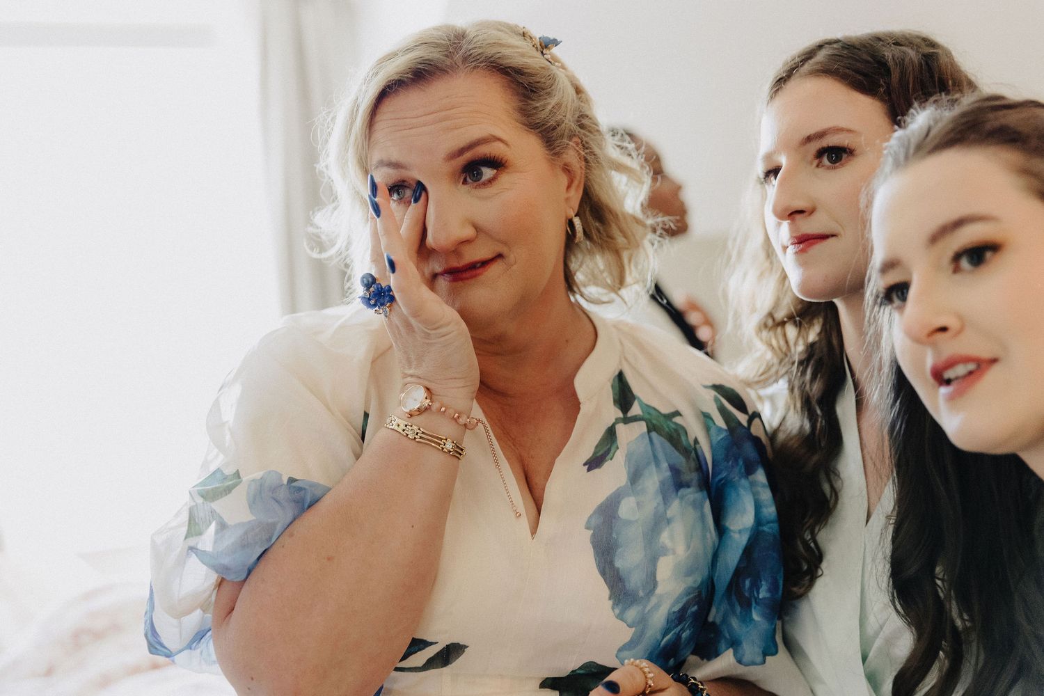 Emotional woman wiping tears at a wedding, flanked by two bridesmaids in white robes during bridal preparations.