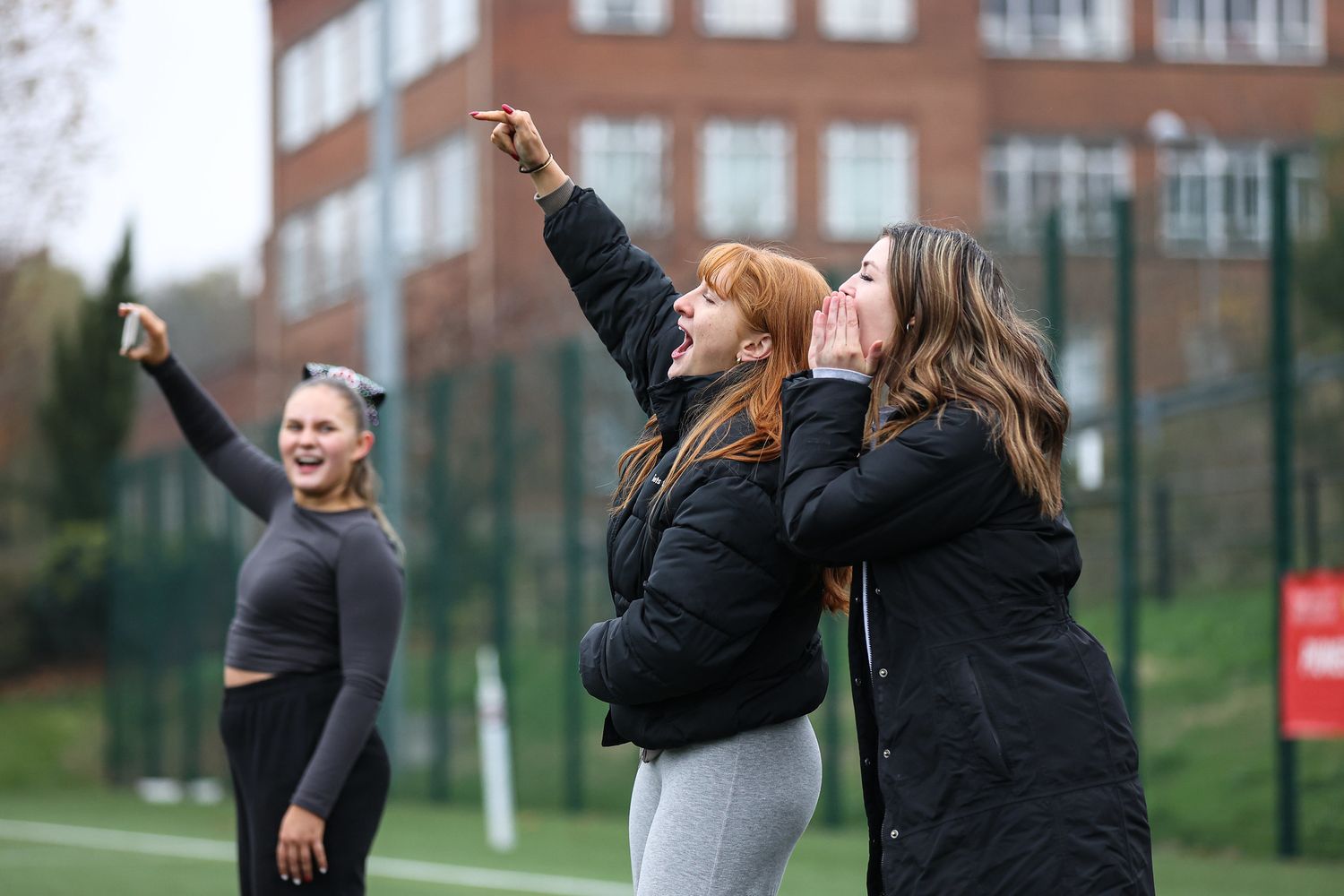 Three people in winter coats enjoying themselves outdoors on a sports field, pointing and looking up at the sky.